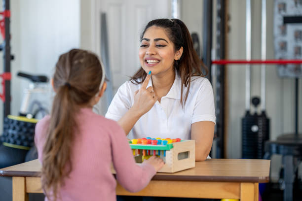 A little girl with a limb difference and a leg brace, sits at a table with her therapist as they work on her speech.  The therapist is using colorful blocks and activities to make the therapy fun.